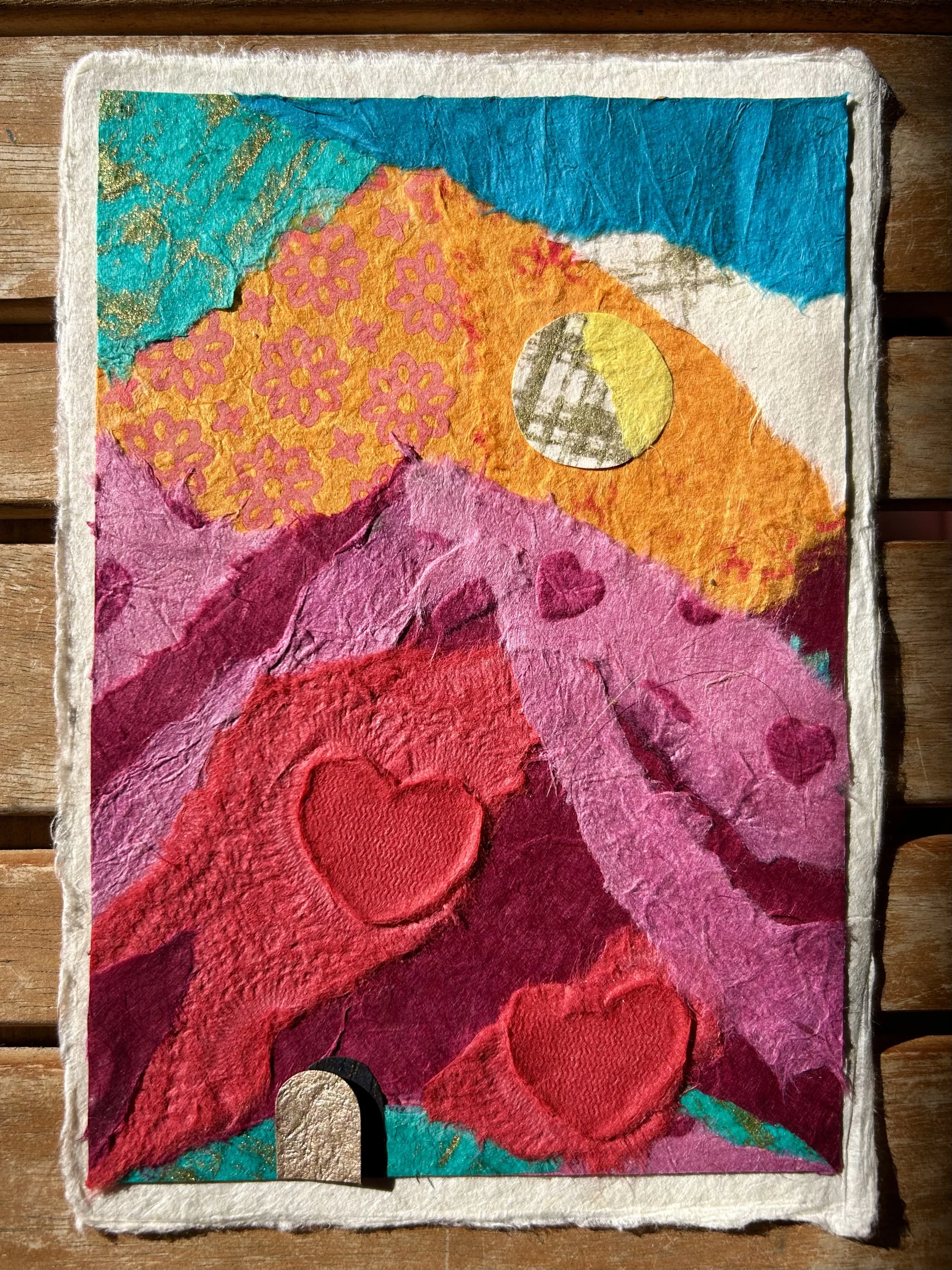 A colorful collage art piece featuring mountains, hearts, a moon, clouds, and a star, made from textured paper and fabric.