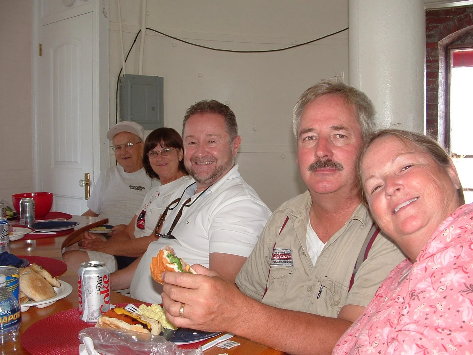 Group of five people sitting at a table with food, soda cans, and sandwiches, smiling in the kitchen of Middle Ground Lighthouse. 