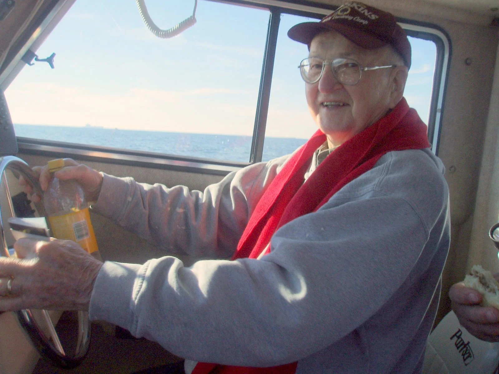 An elderly man with glasses, wearing a red cap and red scarf, sitting in the pilot's seat of a boat, smiling and holding a plastic bottle, with the Chesapeake Bay and sky visible through the window behind him.