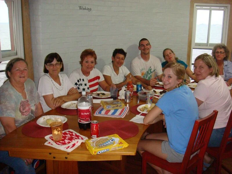 A group of women and a young girl sitting around a dining table with food and drinks, smiling for the photo in the kitchen of Middle Ground Lighthouse. 
