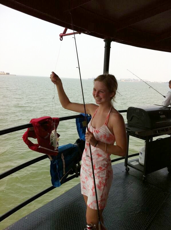 A young woman in a pink and white dress fishing off Middle Ground Lighthouse, smiling. There are fishing rods and a grill on the boat.