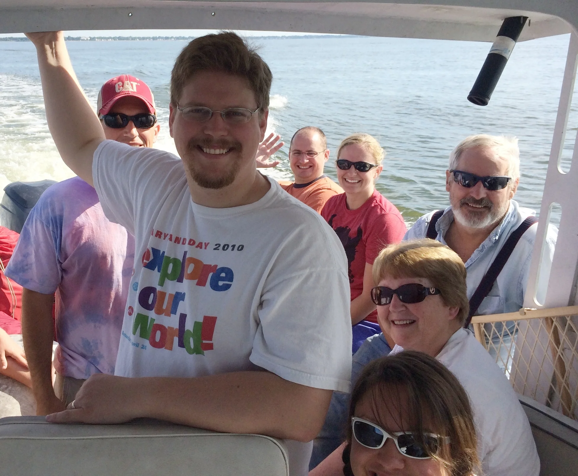 Group of smiling people on a boat, posing for a photo with water and shoreline in the background.