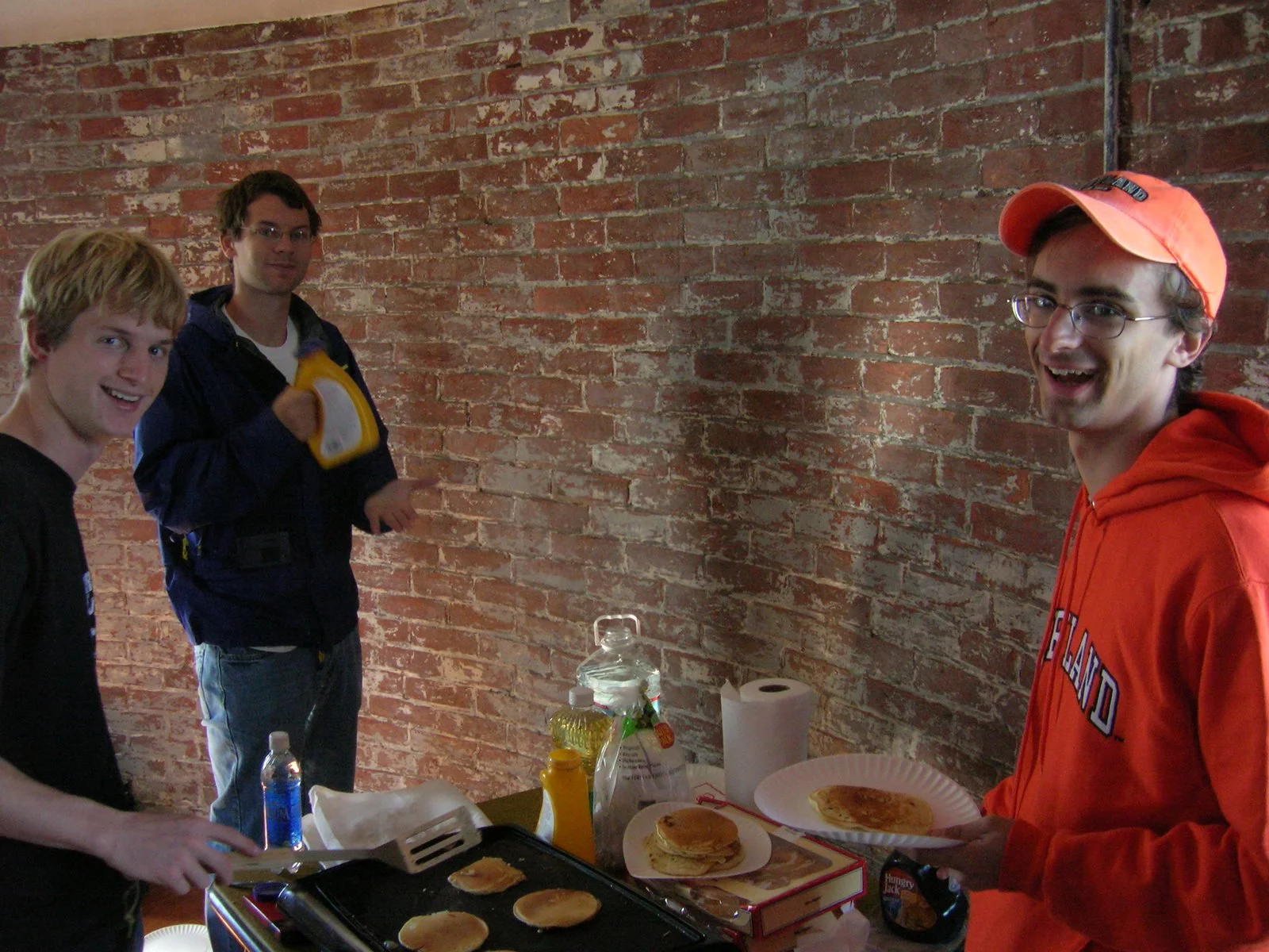 Three young men preparing breakfast on Middle Ground Lighthouse. One is cooking pancakes on a griddle, another holding a spatula, and the third holding a plate of pancakes, smiling.
