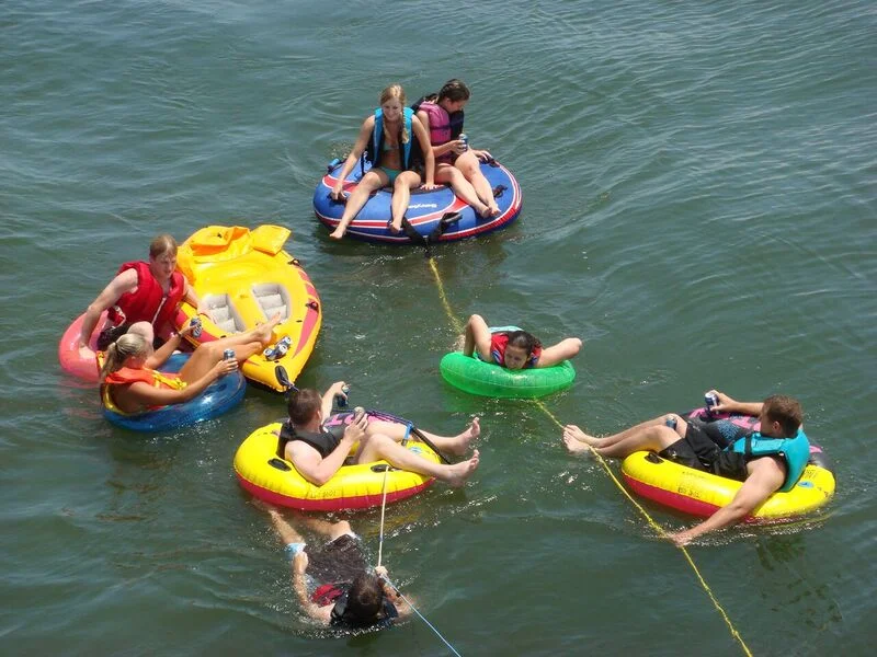 Children and adults on colorful floaties and a raft in the water near Middle Ground Lighthouse, with some using ropes to tow or hold onto the floaties.
