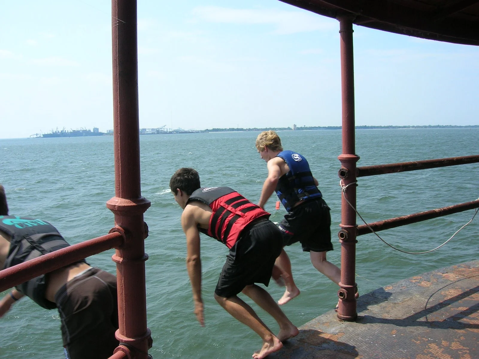 Three boys jumping into the water from the main deck of Middle Ground Lighthouse with a large body of water and a distant shoreline with Hampton Roads in the background.