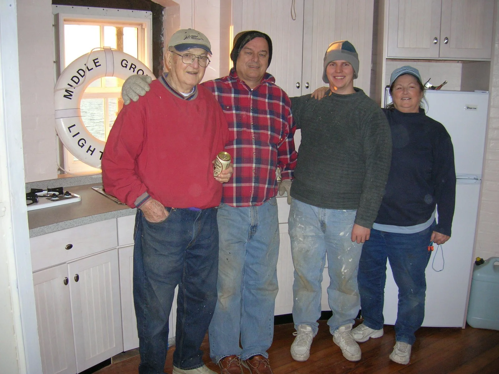 Four people standing together inside a kitchen, smiling at the camera. One person is wearing a red sweater, the others are in casual clothing with beanies or hats. A life preserver with the words 'Middle Grounds Light' is on the wall behind them, and