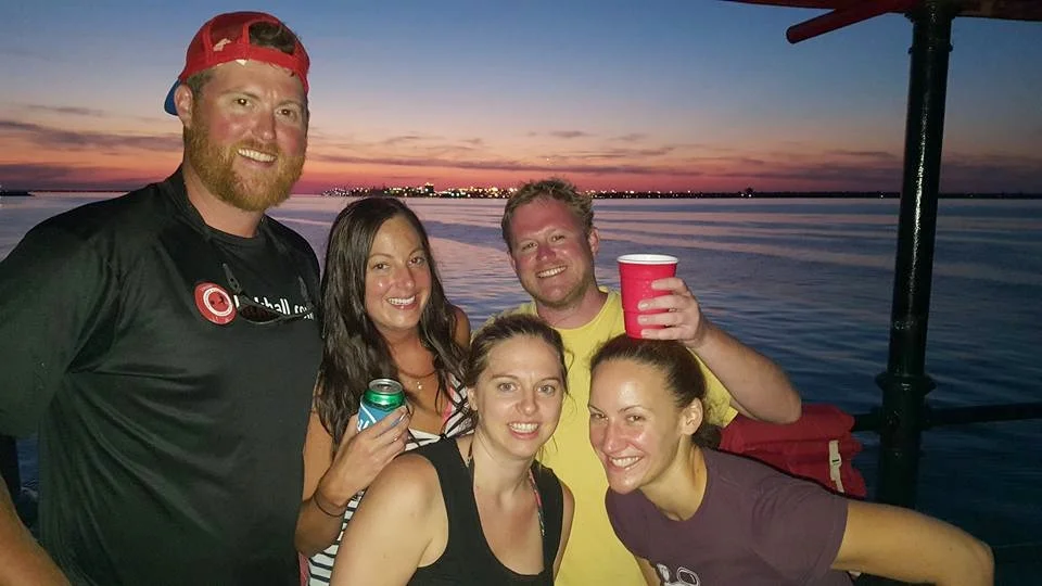 Group of five friends smiling on a boat during sunset, with water and distant city lights in the background.
