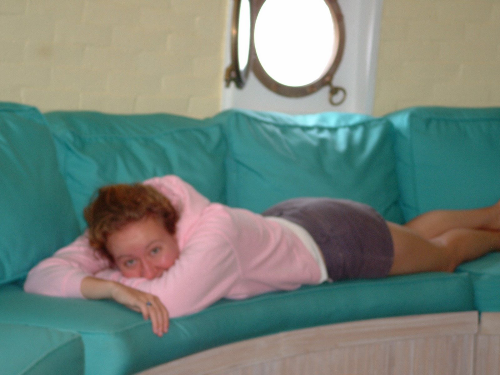 A woman lying on her side on a teal couch, in the porthole room of Middle Ground Lighthouse. There is a porthole window behind her in the background.