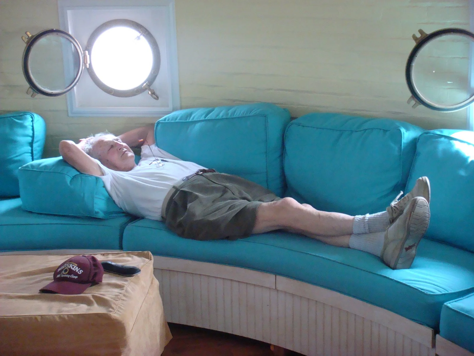An elderly man lying on a large, turquoise sectional sofa with his hands behind his head, relaxing with closed eyes, near two porthole windows, in the porthole room of Middle Ground Lighthouse.