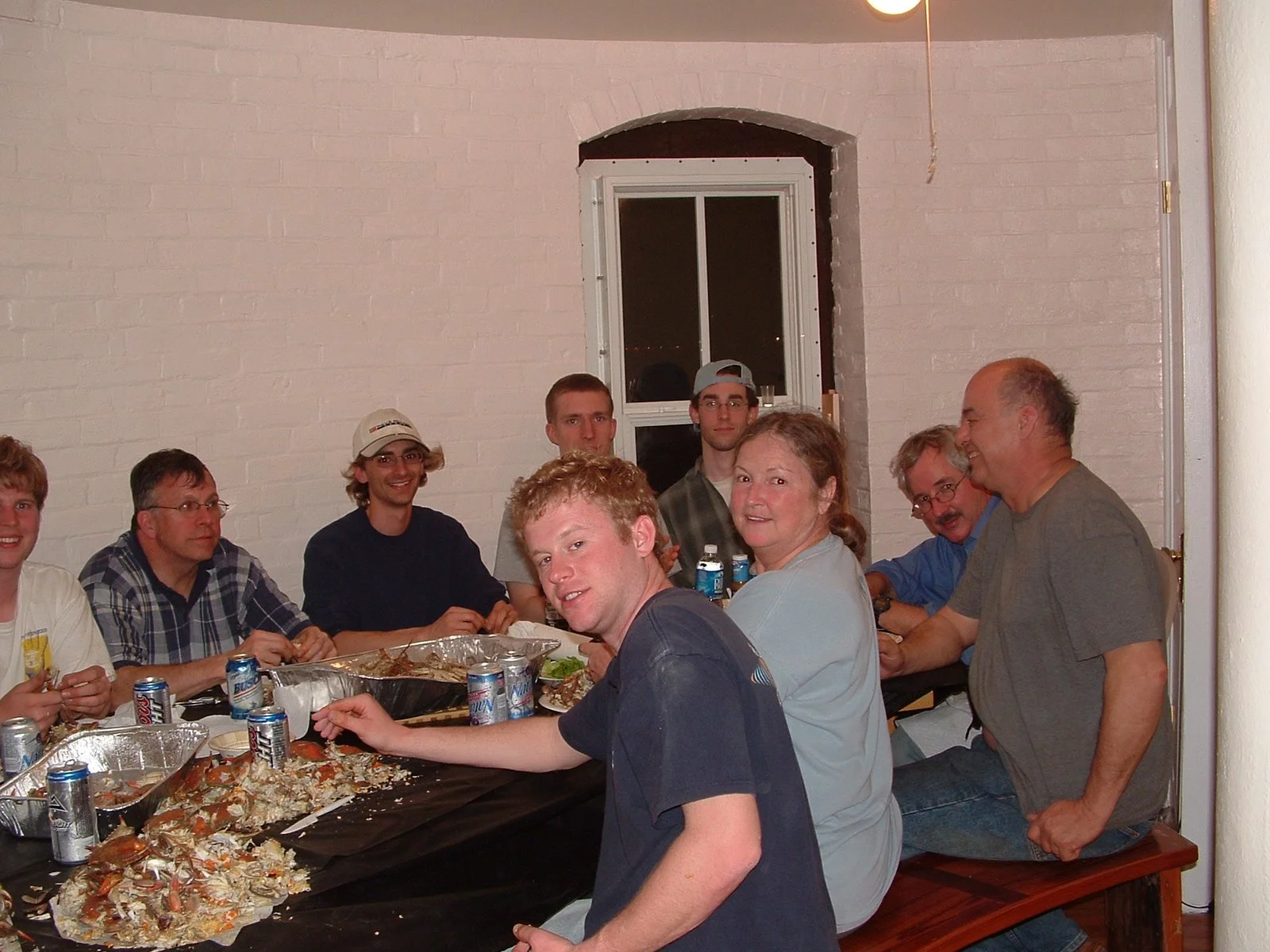 A group of people gathered around a table with leftover food, drinks, and foil trays, celebrating together in the kitchen of Middle Ground Lighthouse. 