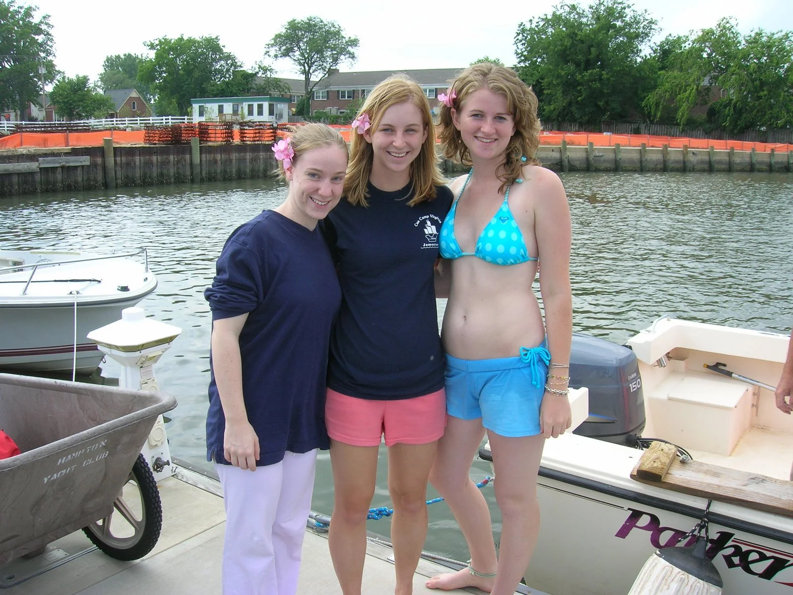 Three young women standing on a dock by the water, smiling at the camera.