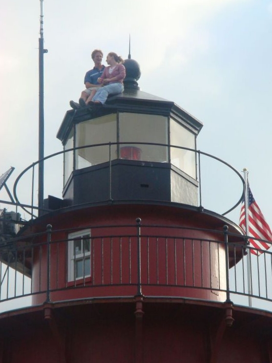 Two people sitting on top of Middle Ground Lighthouse lantern room, with an American flag nearby.