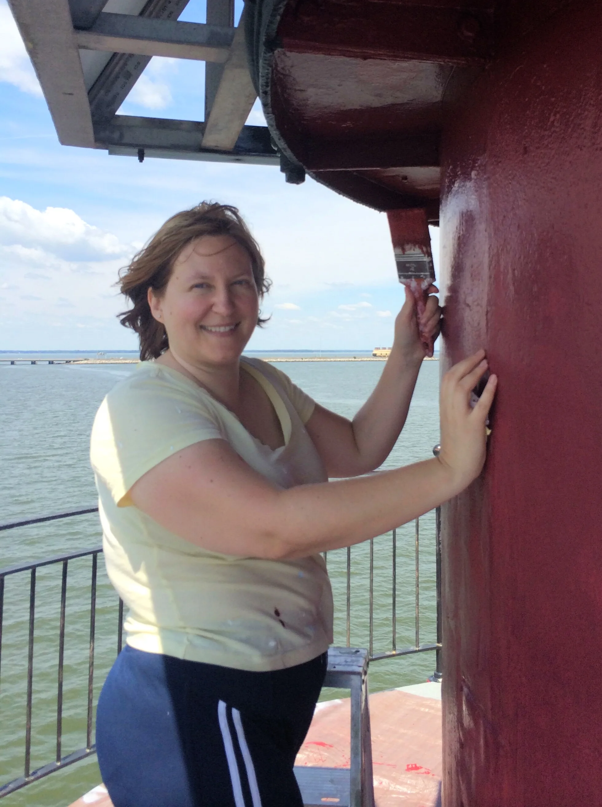 A woman smiling on a boat near a body of water, with sky and clouds in the background, holding onto a wooden structure painted red.