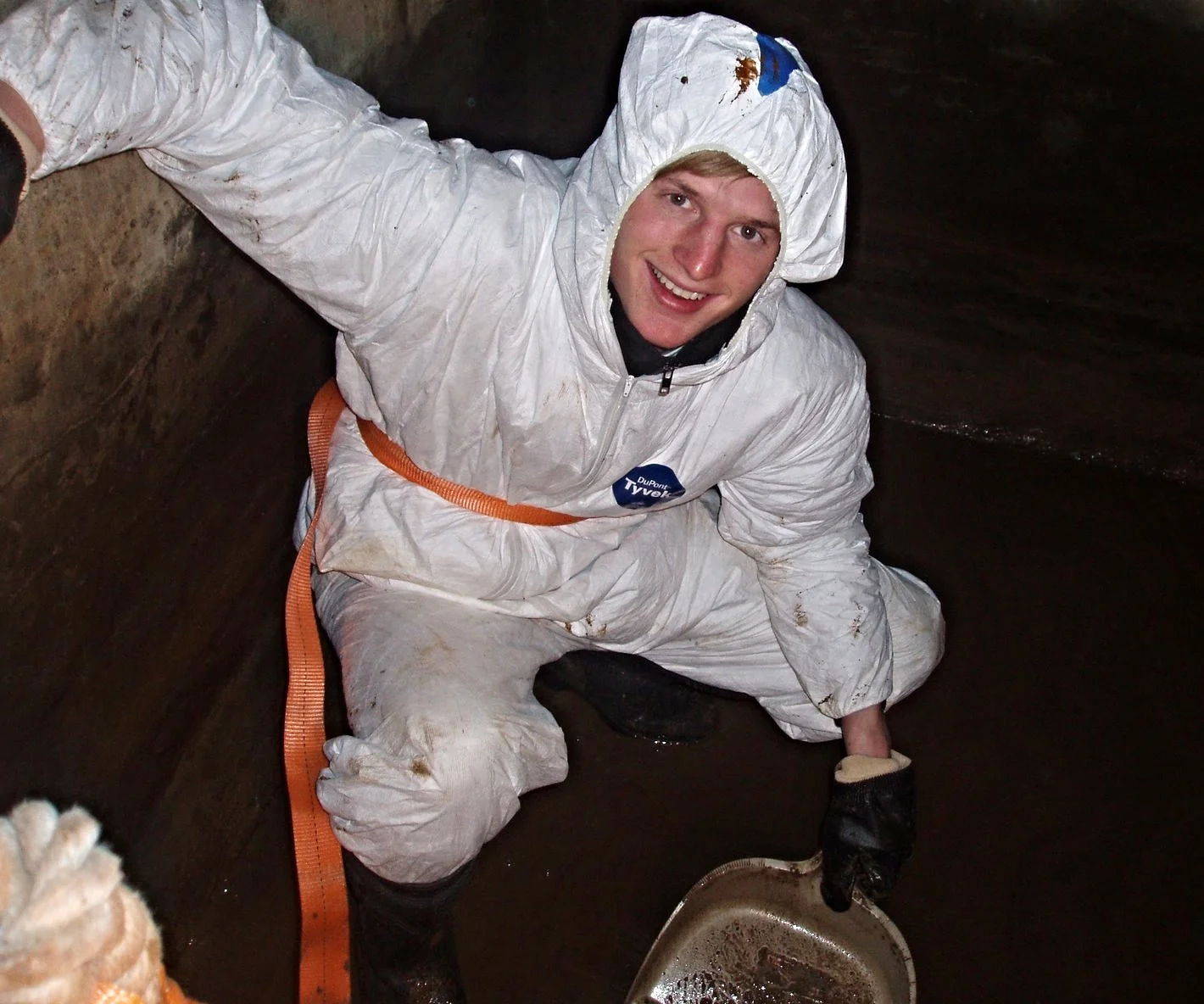 A person wearing a white protective suit, black gloves, and an orange safety harness, crouching and cleaning out the cellar cisterns of Middle Ground Lighthouse. 