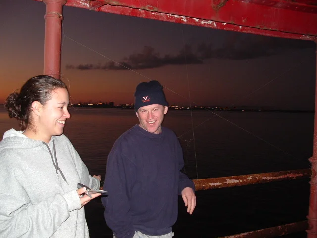 Two people standing on the deck Middle Ground Lighthouse during sunset, with water and a distant horizon of Hampton Roads in the background. 