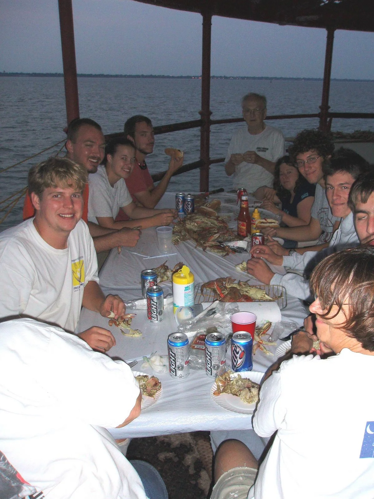 Group of people sitting around a table on the main deck of Middle Ground Lighthouse, enjoying a seafood meal with cans of beer, hot dogs, and drinks, with a water body in the background during dusk.