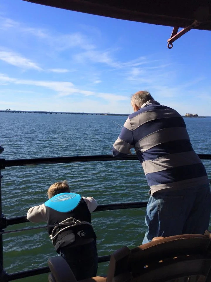 A man and a young boy fishing off Middle Ground Lighthouse, looking out over calm water with the Monitor-Merimac Bridge-Tunnel and clear blue sky in the background.