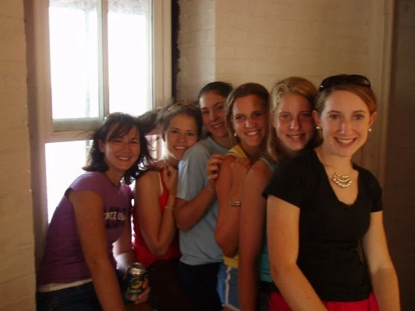 Six young women standing in a line indoors near a window, smiling at the camera in the kitchen of Middle Ground Lighthouse. 