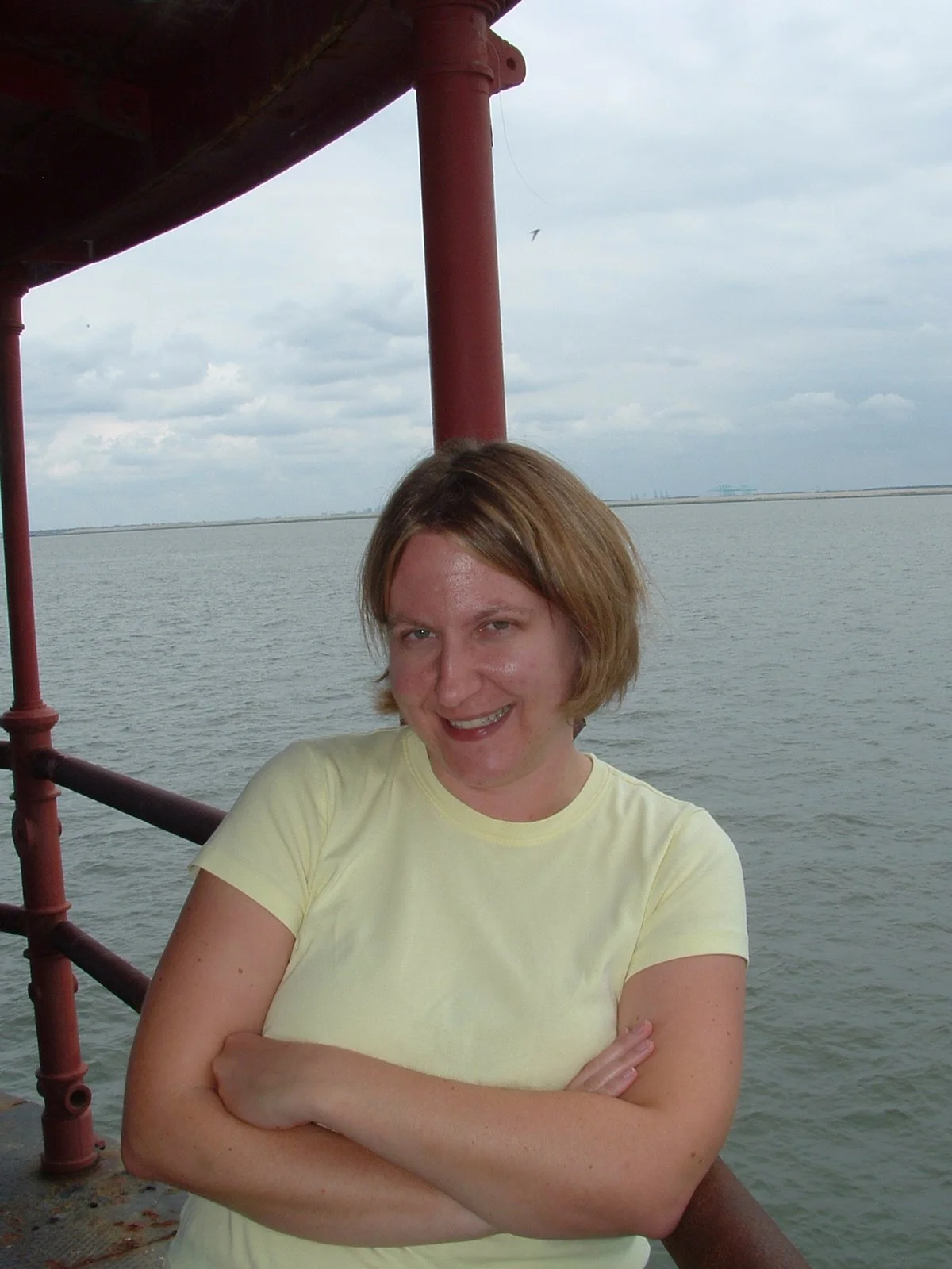 A woman with short light brown hair wearing a yellow shirt standing on the main deck of Middle Ground Lighthouse with water and cloudy sky in the background.