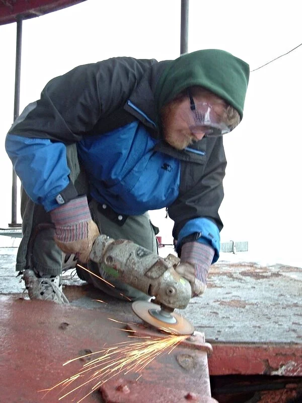 A man wearing safety goggles cutting metal with an angle grinder, creating sparks, while restoring the main deck of Middle Ground Lighthouse.
