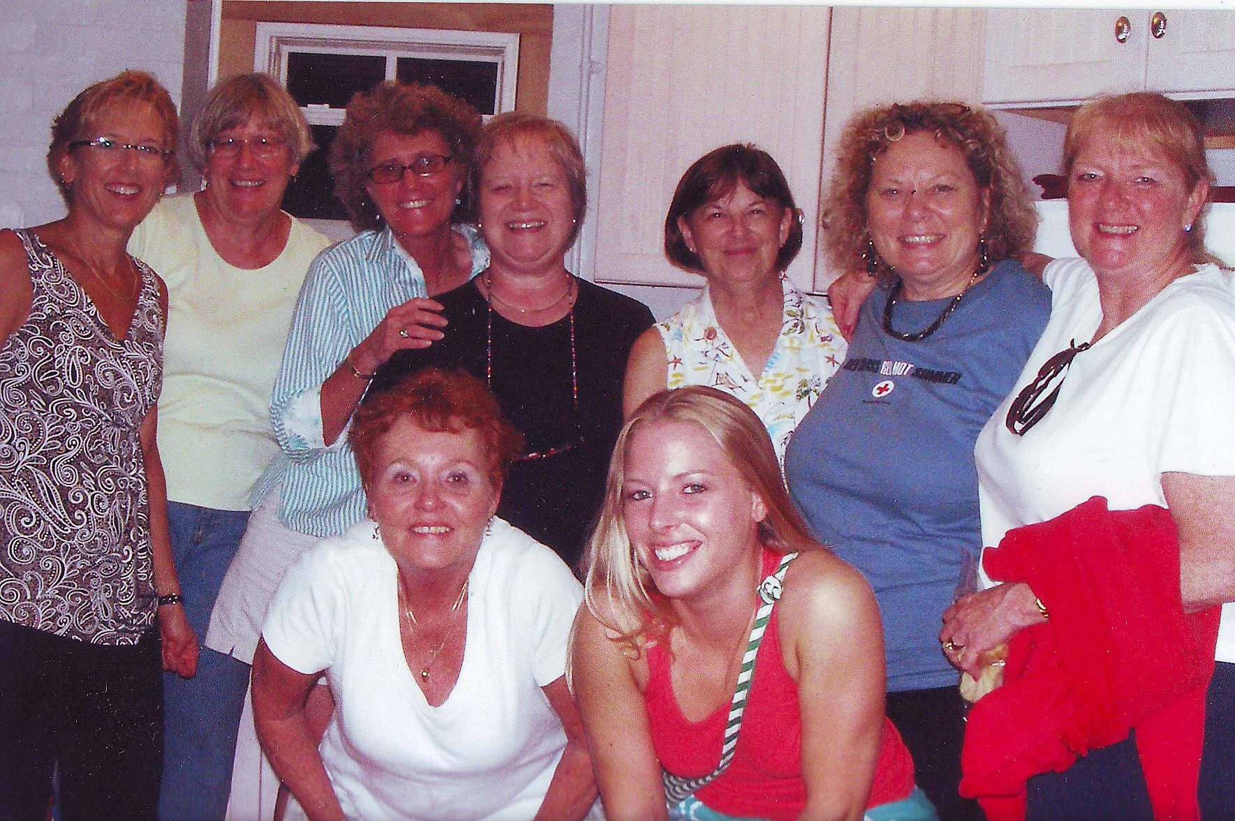 Group of women gathered together, smiling, in the kitchen of Middle Ground Lighthouse. Some women are standing while others are seated, and they are wearing casual summer clothing.
