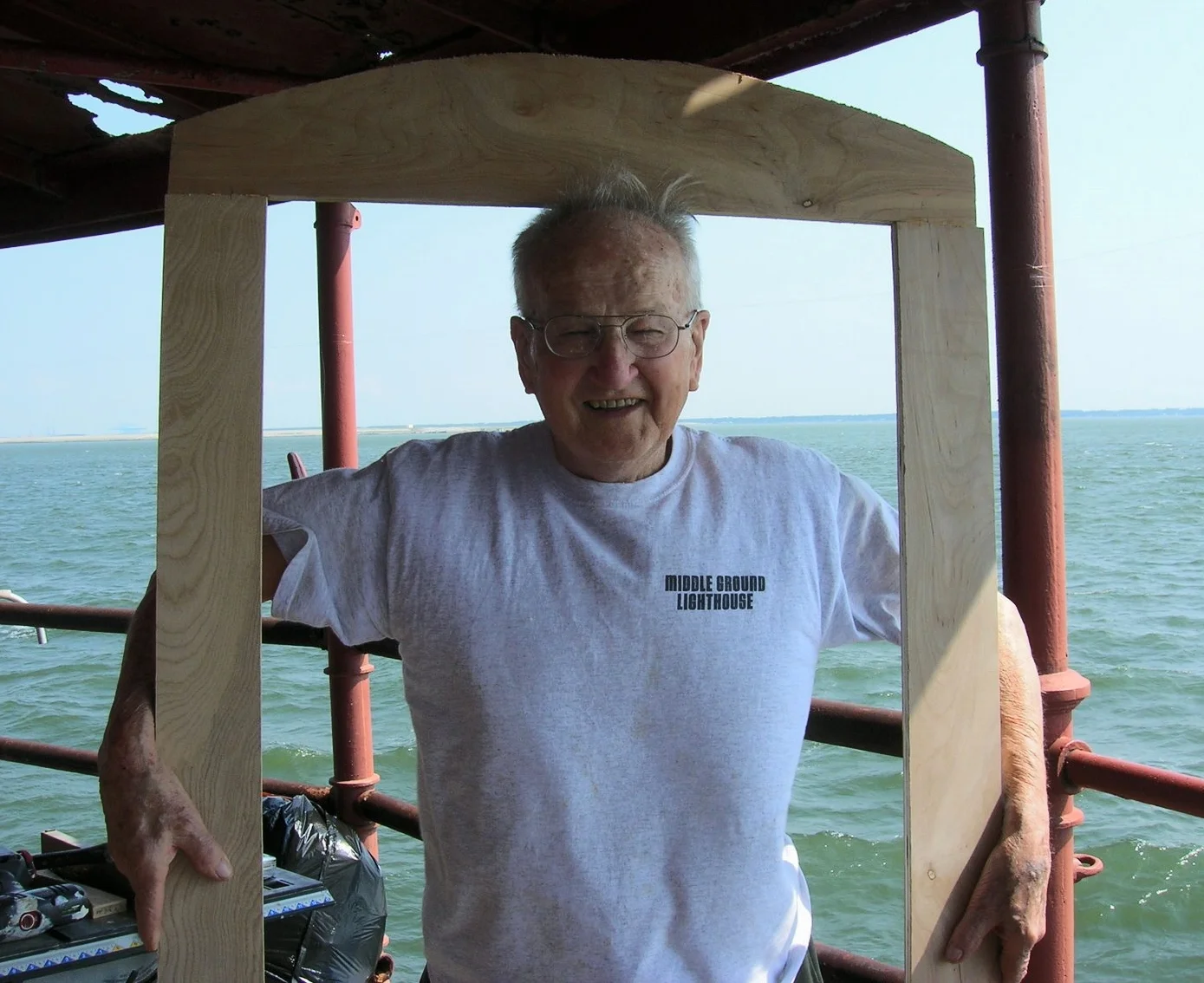 An elderly man with glasses and a white t-shirt labeled 'Middle Ground Lighthouse' is smiling, standing on a boat with a wooden frame around him, water and a distant shoreline in the background.