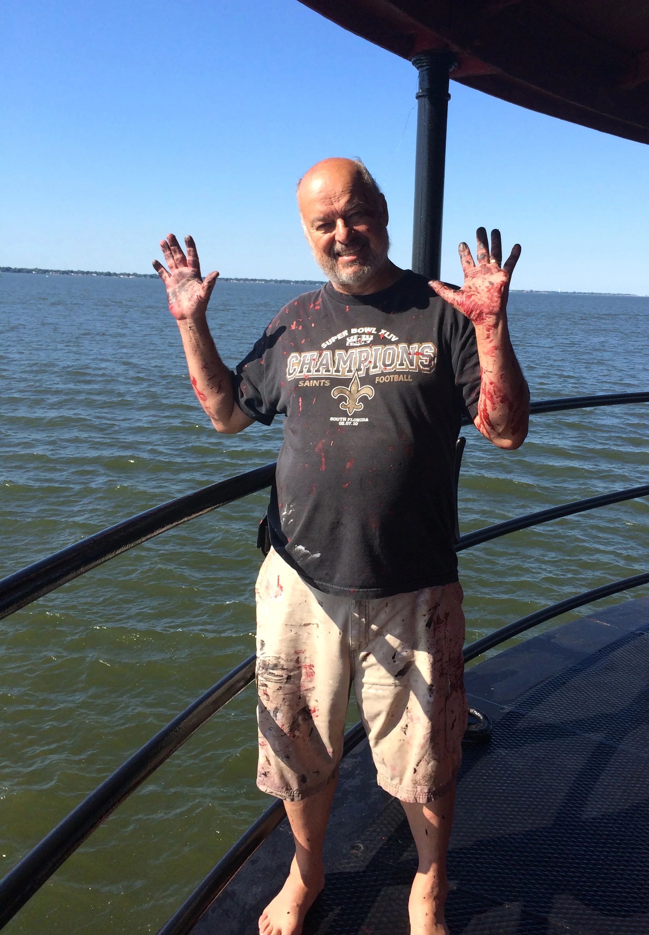 A man standing on Middle Ground Lighthouse by the water, smiling and waving with his hands covered in paint. 