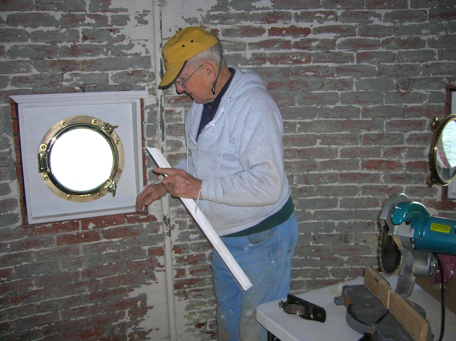 An older man working to install new porthole windows in the porthole room of Middle Ground Lighthouse. There is a table with tools nearby, including a power saw and a clamp.