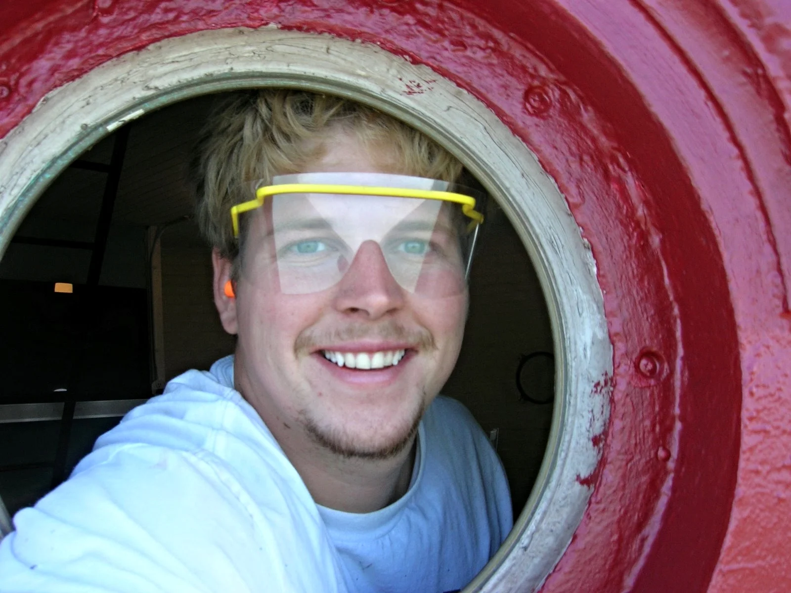 A young worker smiling while taking a selfie through a porthole on Middle Ground Lighthouse, wearing safety goggles.
