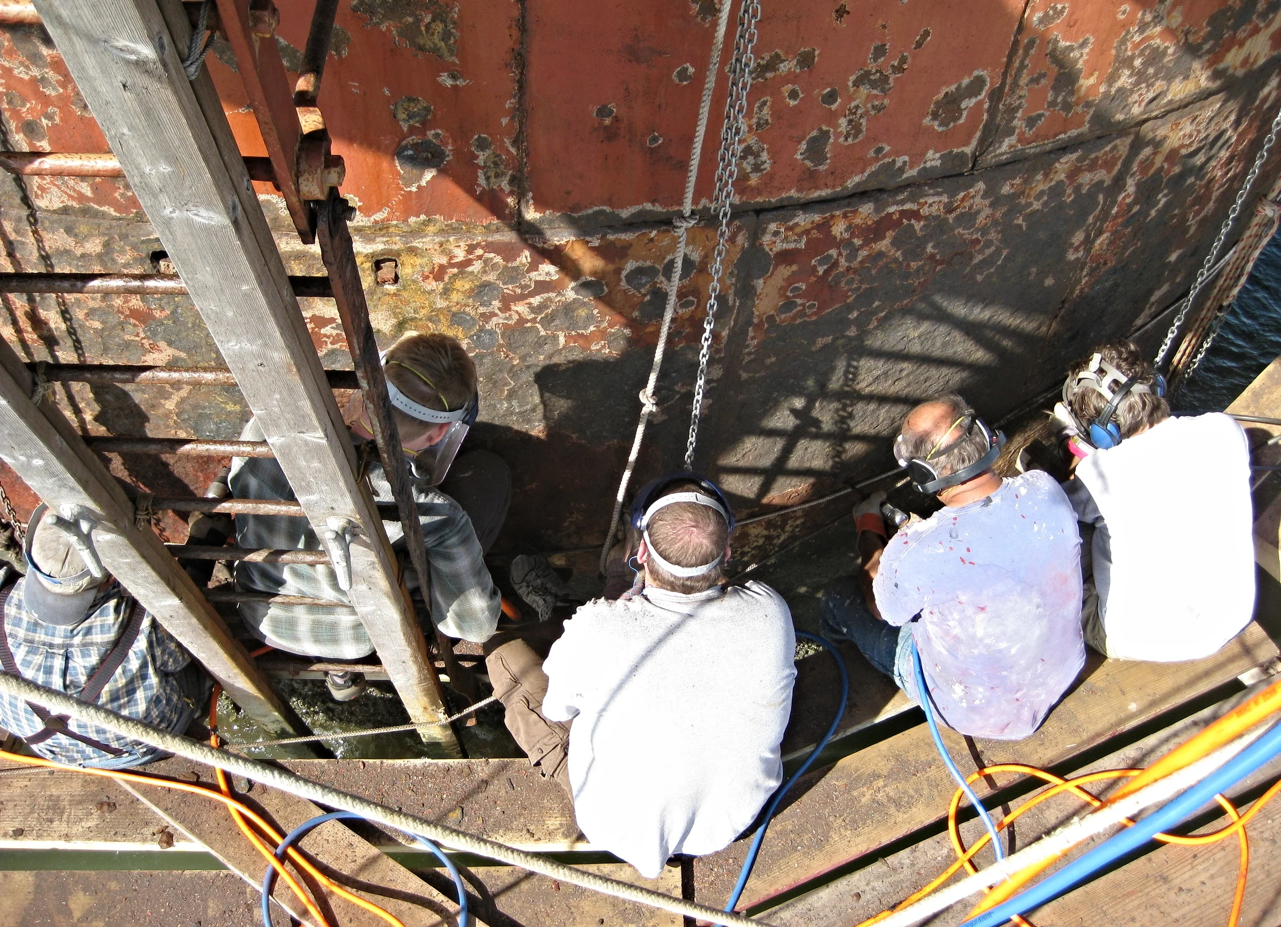 People working on the exterior of Middle Ground Lighthouse from a scaffolding platform, wearing protective gear while inspecting the rusty, weathered surface.