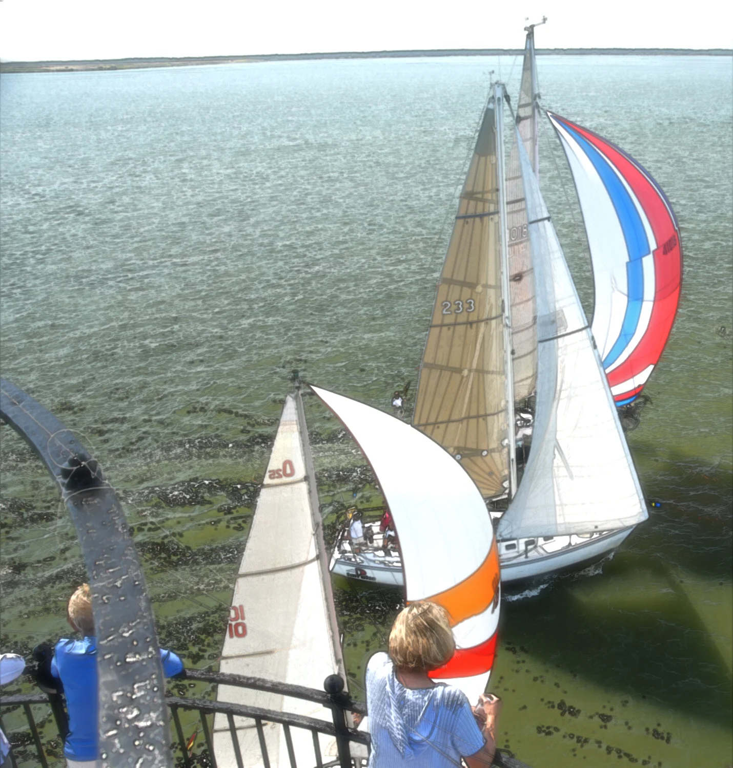 Multiple sailing boats with colorful sails surrounding Middle Ground Lighthouse, with people onboard and a view from a pier or dock in the foreground.