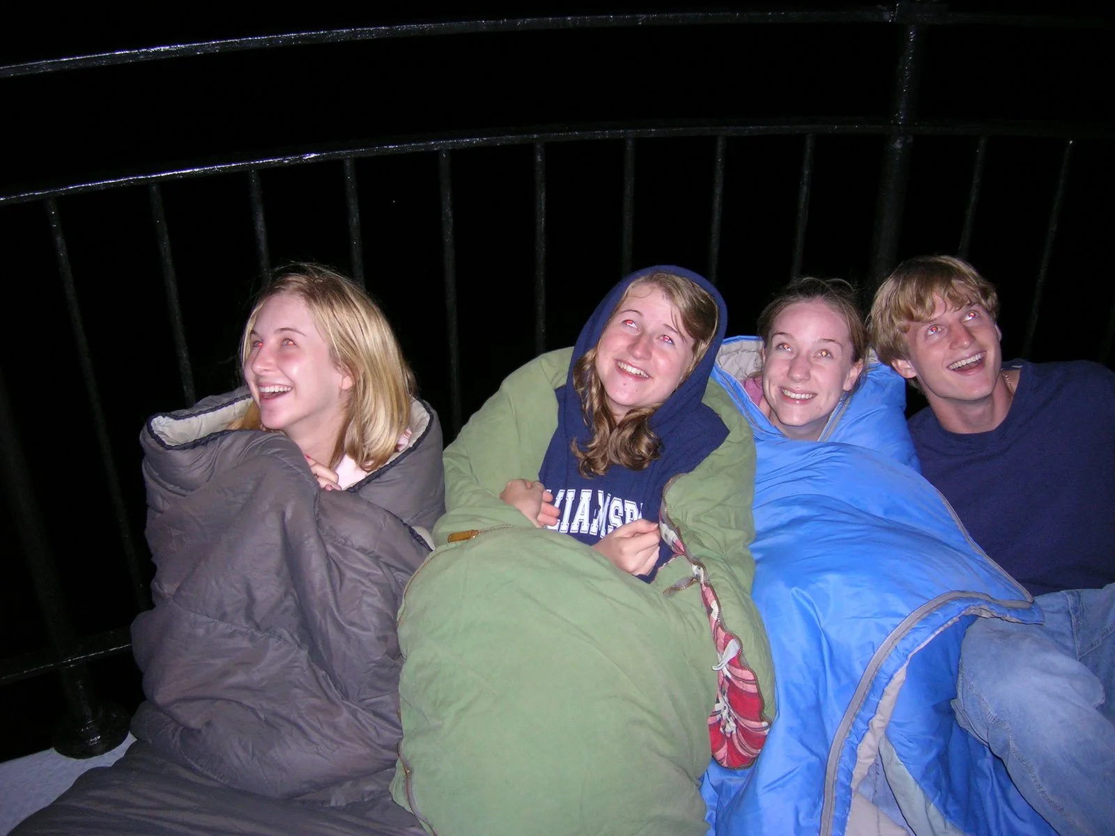 Four teenagers lying on sleeping bags outdoors at night, smiling and looking up at fireworks over Middle Ground Lighthouse. 