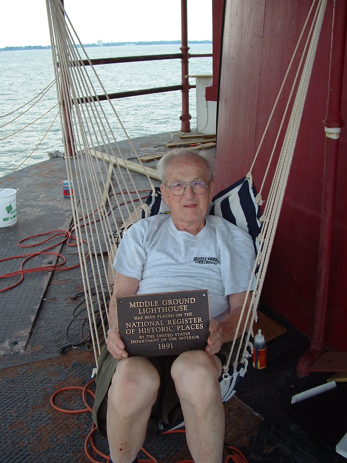 An elderly man sitting in a hammock, holding a plaque that states the Middle Ground Lighthouse is on the National Register of Historic Places, with water and a distant shoreline in the background.