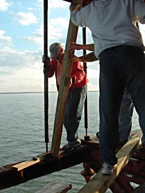 Workers installing new deck materials on the main deck of Middle Ground Lighthouse, with a sunset sky in the background.