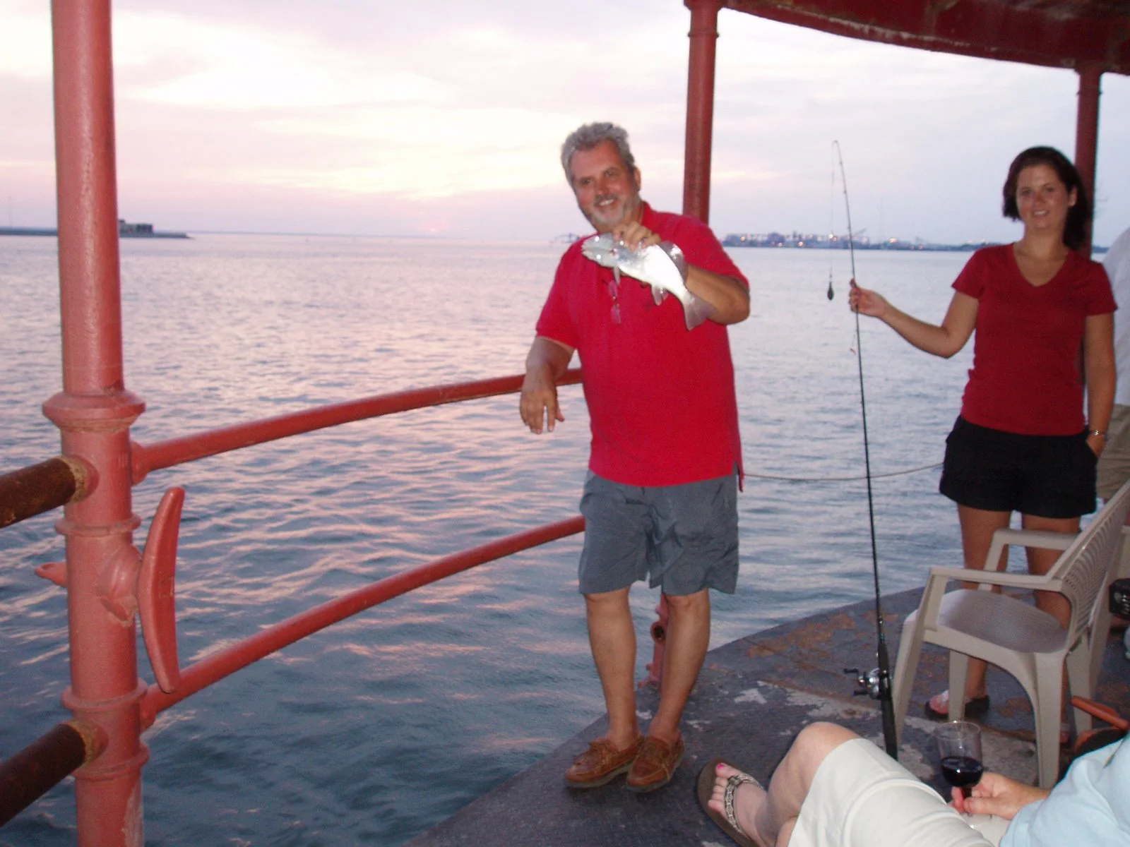 A man in a red shirt holding a fish on the main deck of Middle Ground Lighthouse during sunset with two women nearby, one with a fishing pole, in a body of water.