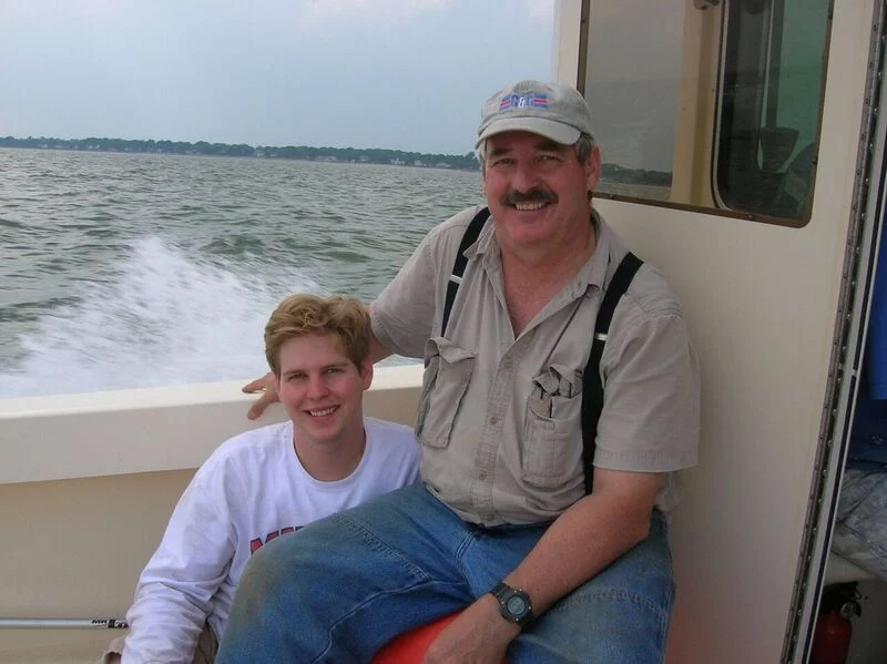 A smiling man and a young man sitting on a boat with water and a shoreline in the background.