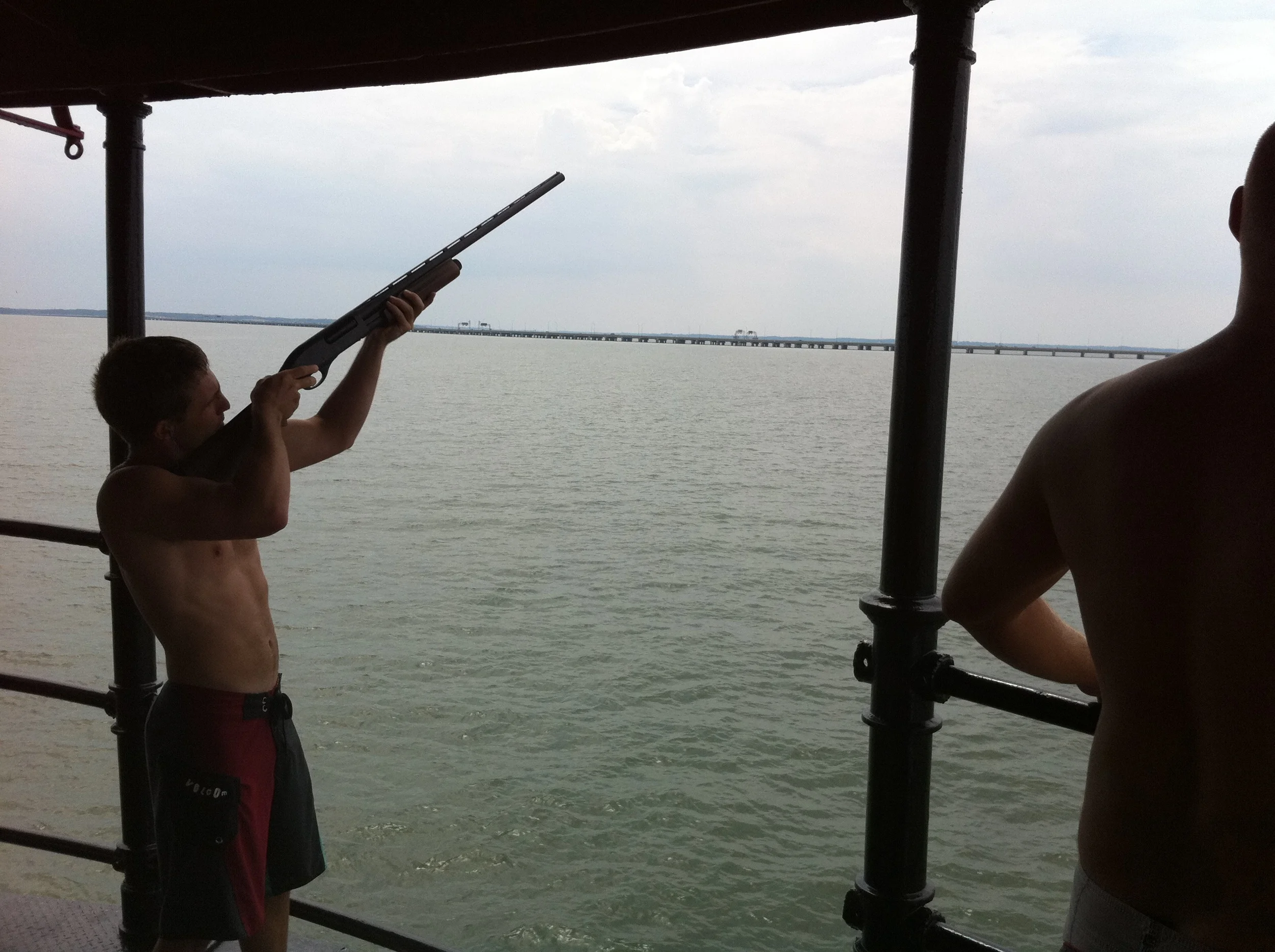 Two shirtless young men skeet shooting off of the main deck of Middle Ground Lighthouse. 