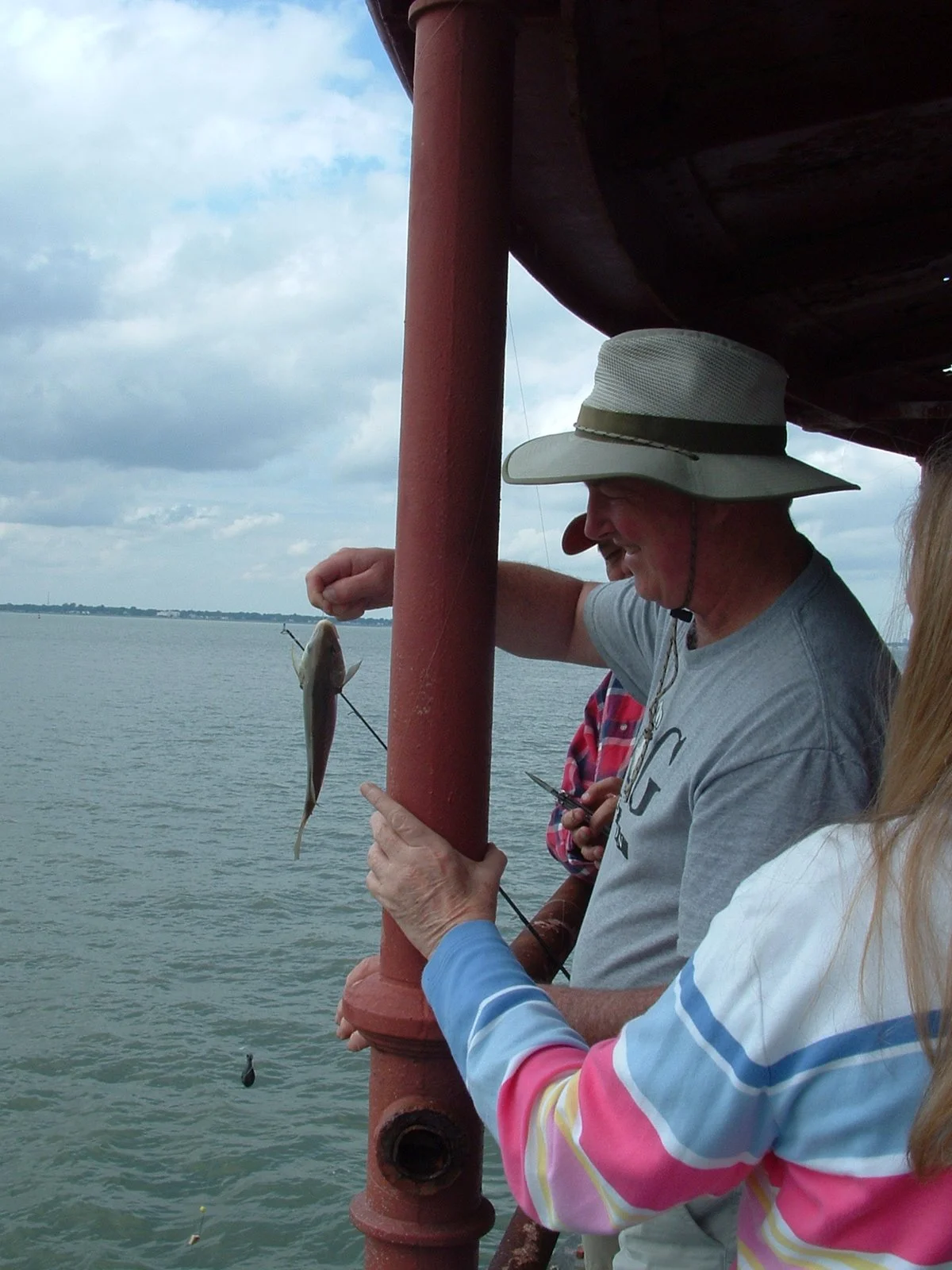 A group of people fishing off Middle Ground Lighthouse, with a calm body of water in the background and cloudy skies overhead.
