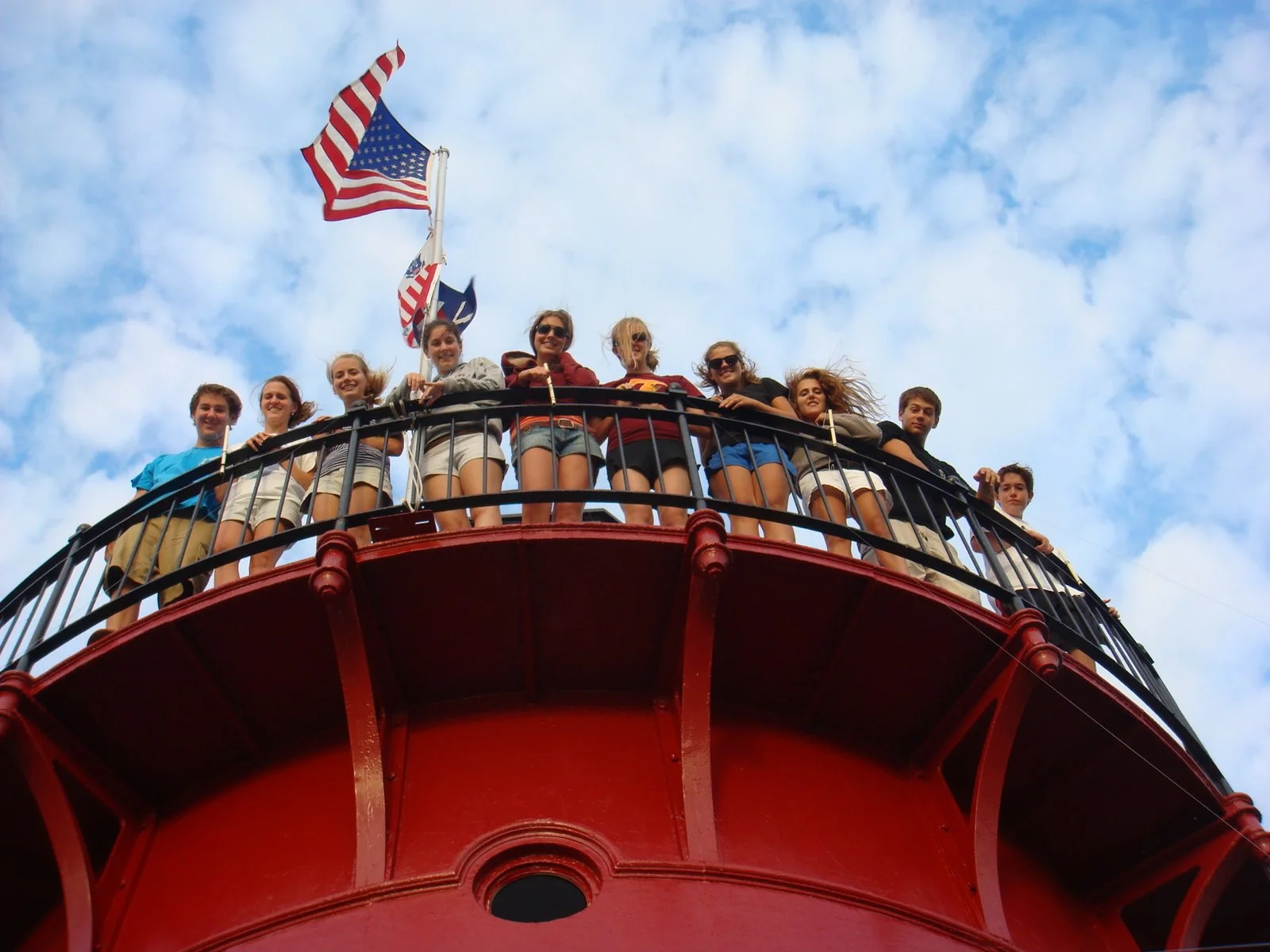 Young people standing on the watch level balcony of Middle Ground Lighthouse, looking down at the camera with flags flying in the background and a partly cloudy sky.