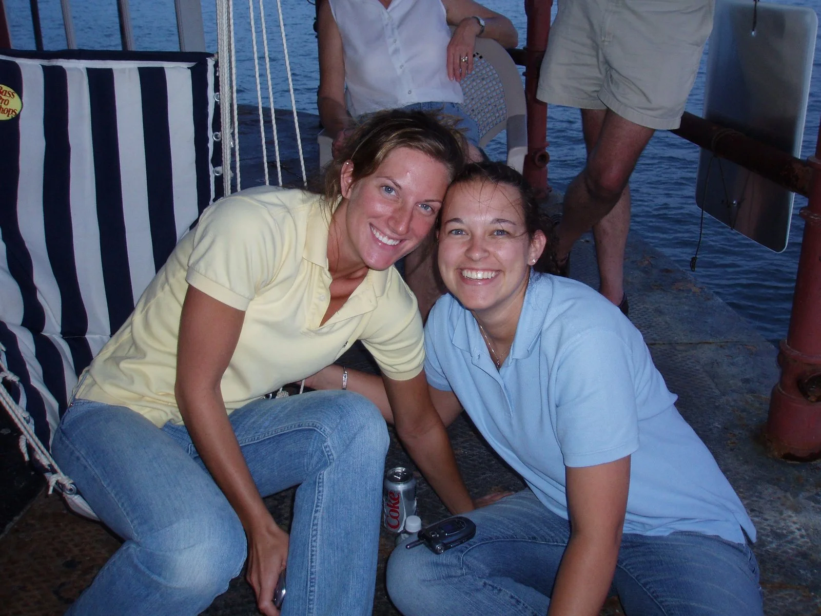 Two young women are sitting on the deck of Middle Ground Lighthouse, smiling at the camera. There is water in the background and parts of other people on the boat are visible.