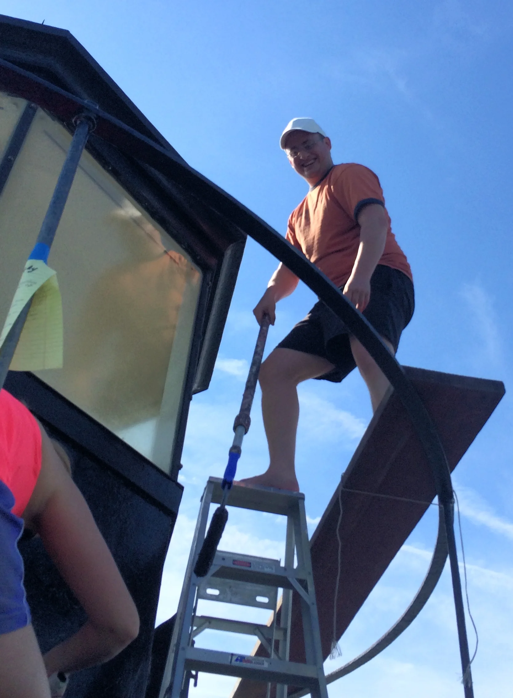 A person standing on a platform at the lantern room level of Middle Ground Lighthouse, smiling at the camera, with a ladder and a frame around them, against a blue sky with scattered clouds.