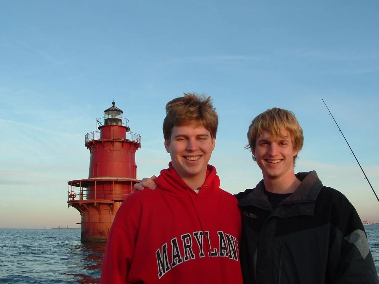 Two young men smiling with their arms around each other by Middle Ground Lighthouse near the water on a clear day.