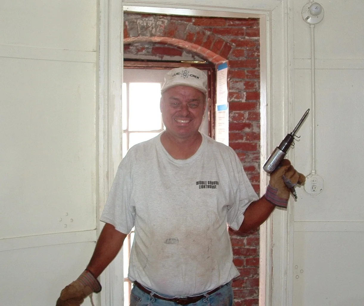 A smiling worker wearing work gloves and holding a caulking gun, standing on Middle Ground Lighthouse. 