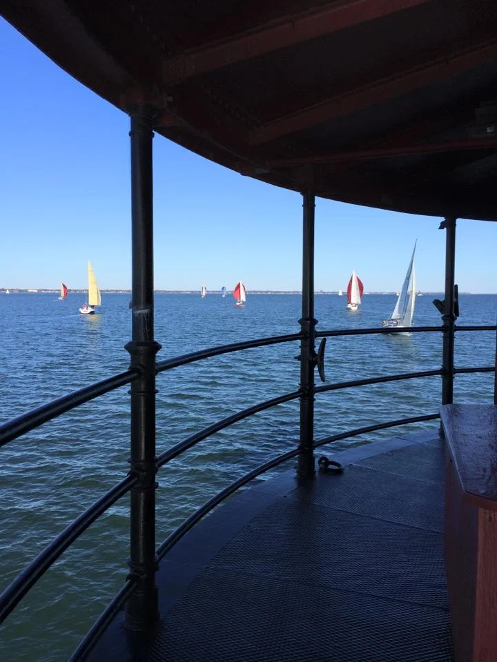 View from the main deck of Middle Ground Lighthouse looking out at sailboats on the water with colorful sails under a clear blue sky.