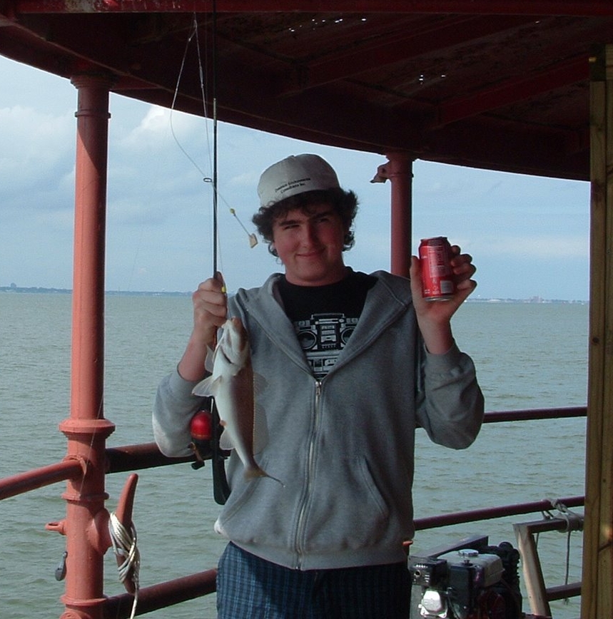 A young man on a boat holding a fish in one hand and a soda can in the other, smiling on the main deck of Middle Ground Lighthouse.