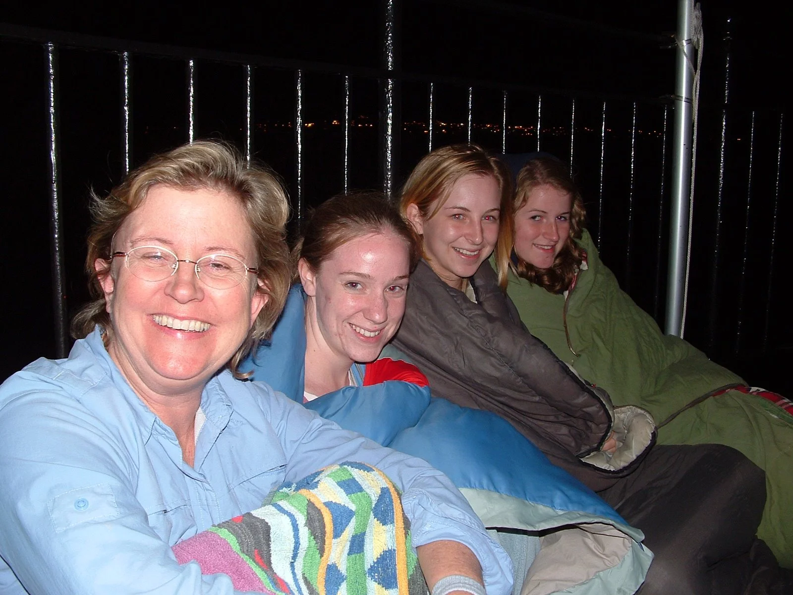 Five women sitting on Middle Ground Lighthouse at night with metal railings behind them, smiling at the camera.