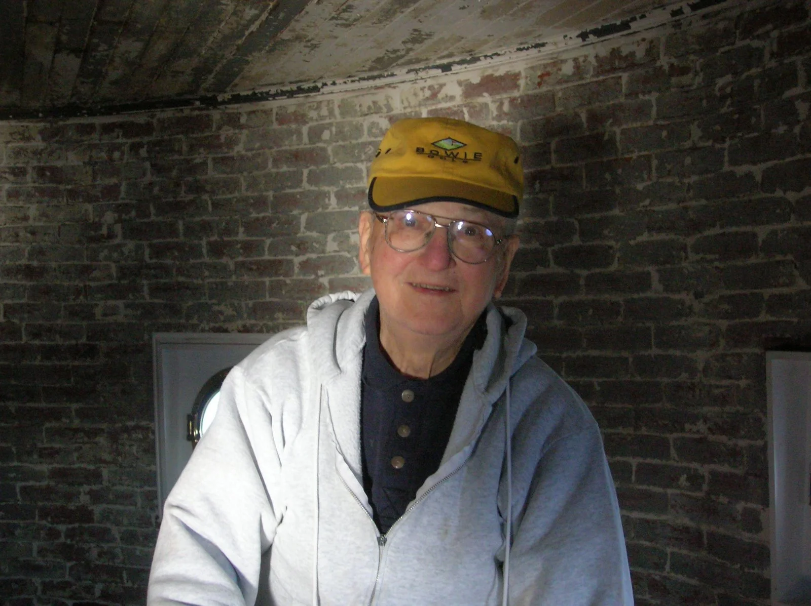 An older man smiling at the camera, with a brick wall background on Middle Ground Lighthouse.
