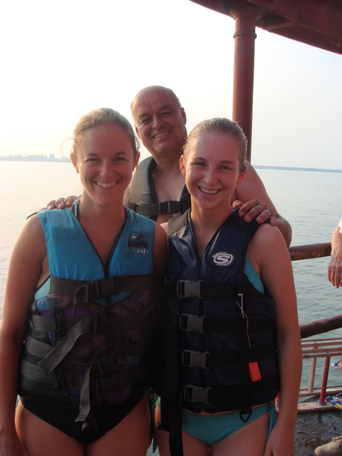Three young people wearing life jackets on Middle Ground Lighthouse, smiling, with water and a distant shoreline in the background.