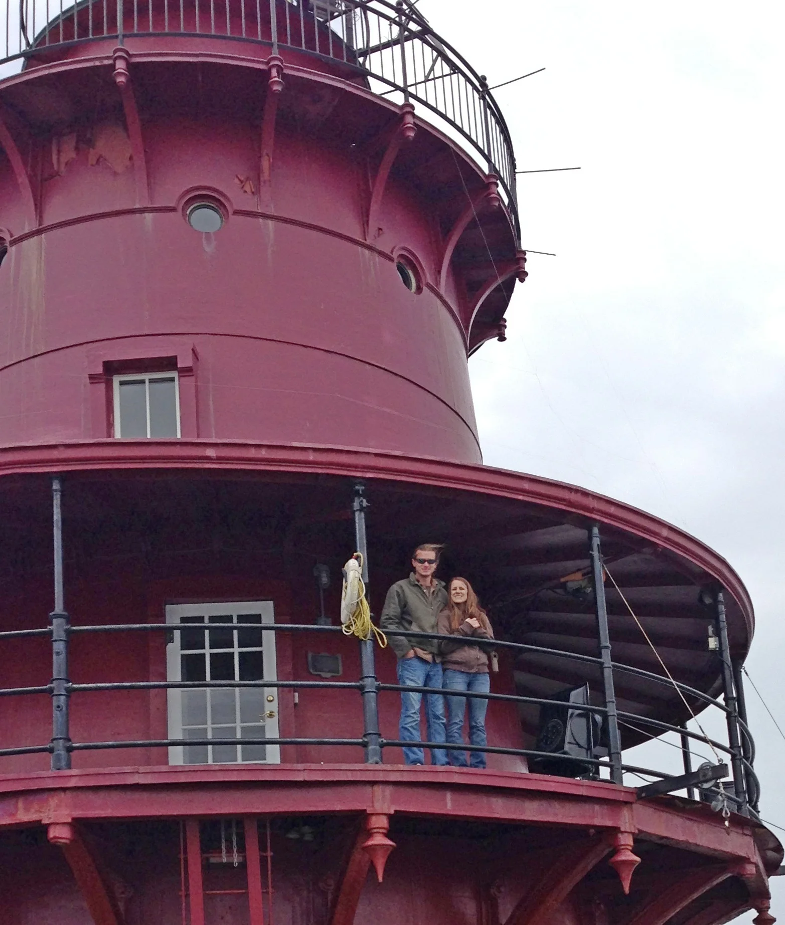 Two people standing on the balcony of Middle Ground Lighthouse smiling at the camera. The lighthouse has multiple levels with railings, windows, and a door visible.
