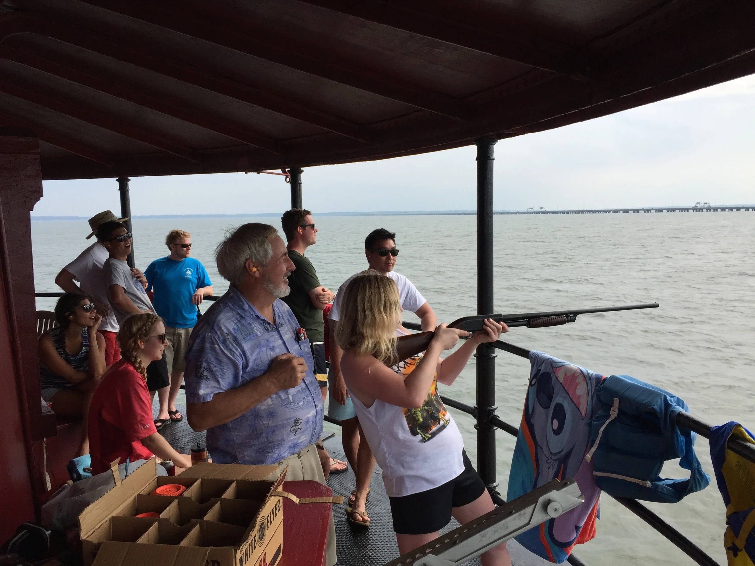 People on a boat from different age groups, including children, teenagers, and adults, looking out over the water, with the Monitor–Merrimac Memorial Bridge–Tunnel visible in the background.